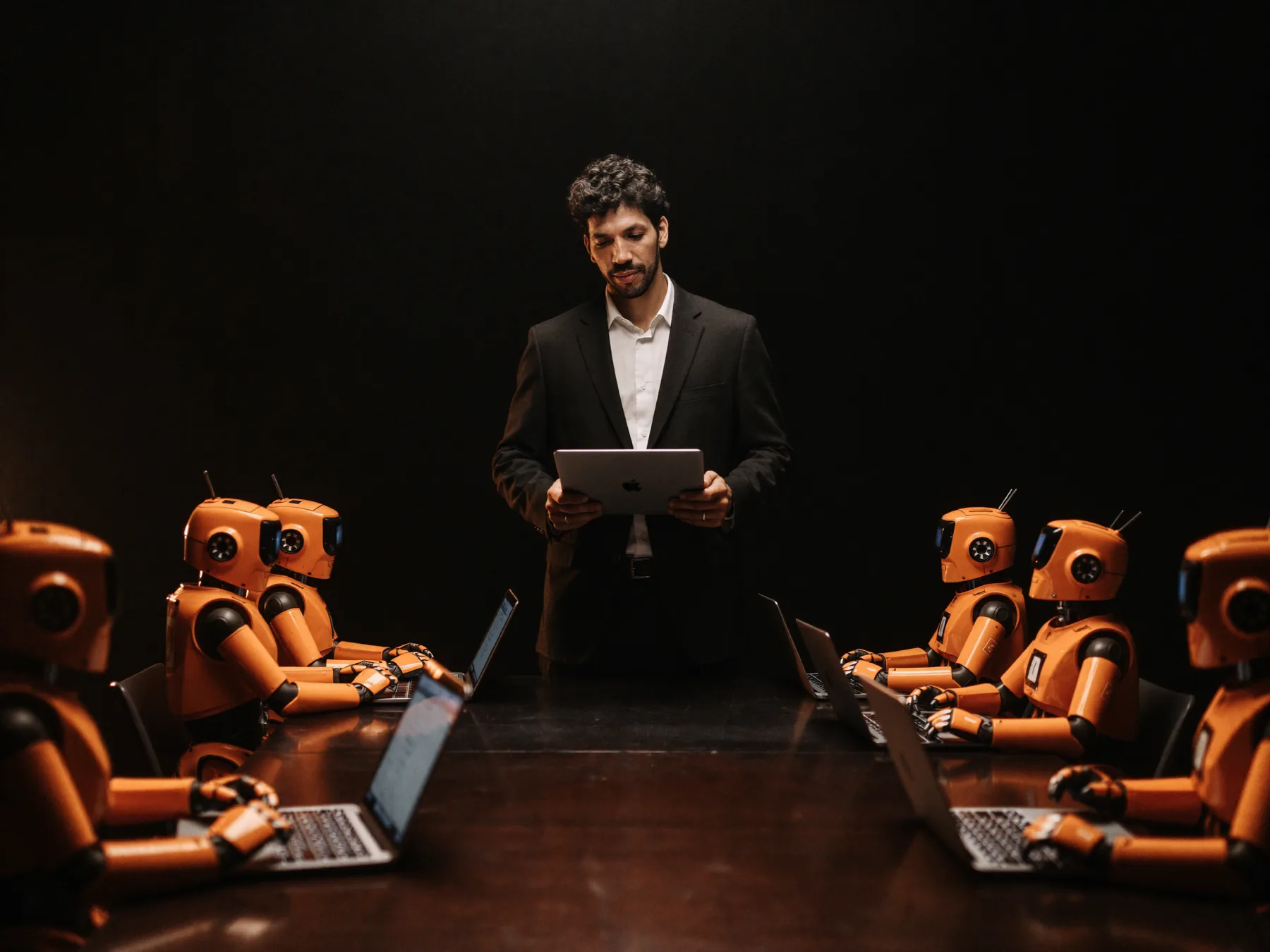 Thales Laray de pé numa mesa escura, orientando um time de robôs laranja com laptops